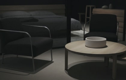 A bedrom focusing a beige table with a bow on top of it and a black chair on the left, the bed is in the back.