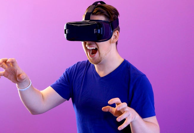 Image of a white man with a VR, wearing a blue shirt, immitating a monster scaring someone with his arms.